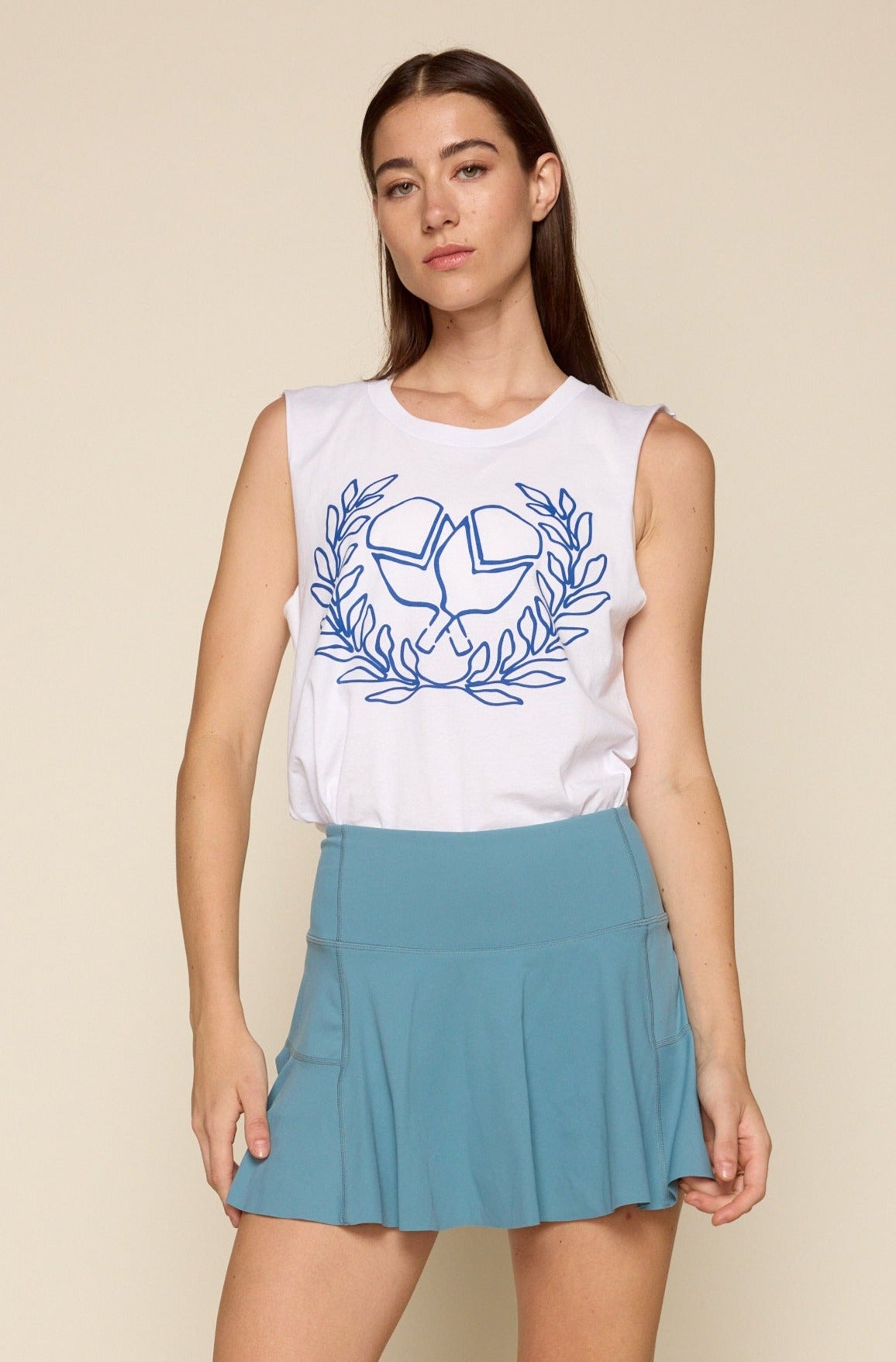 Cropped frontal photo of female model wearing a white muscle tank. Print on the muscle is criss cross pickle ball paddles in blue font surrounded by victory wreath. Model is also wearing a pleated blue skirt