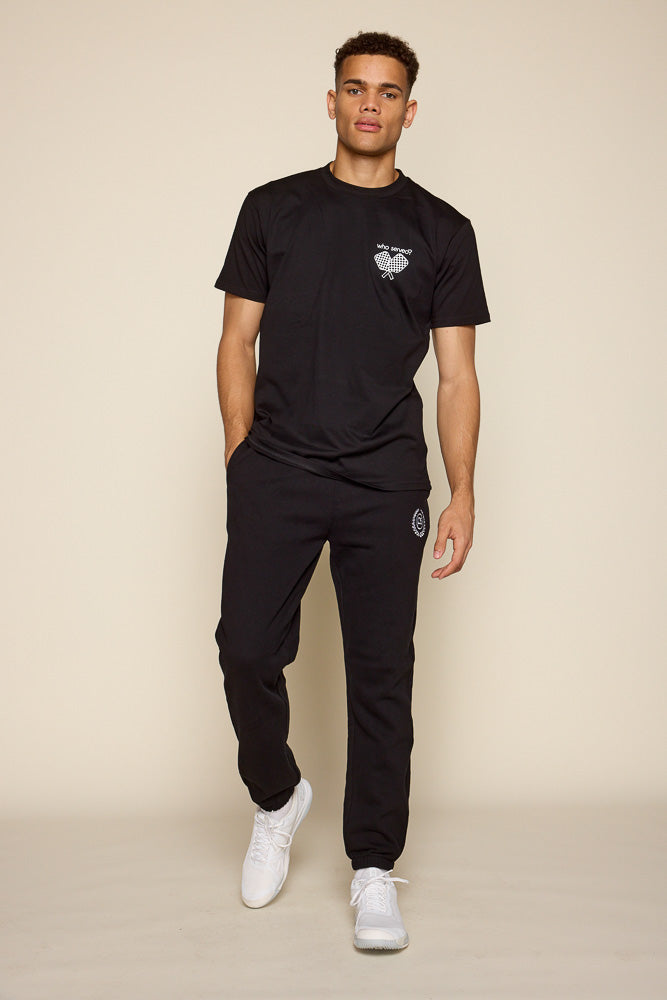 Full frontal view of male model wearing black short sleeve tee. Graphic on the tee is on the upper left chest area in white are two pickleball paddles crisscrossed under the font: "who served?". Model is also shown wearing a black pair of joggers and white sneakers.