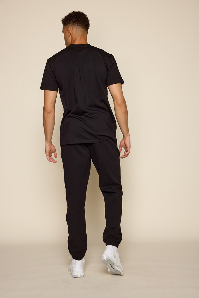 Full back view of male model wearing a black short sleeve tee. Model is also wearing a pair of black joggers and white tennis shoes