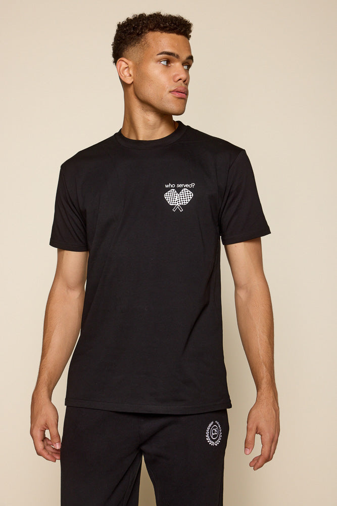 frontal view of male model wearing black short sleeve tee. Graphic on the tee is on the upper left chest area in white are two pickleball paddles crisscrossed under the font: "who served?"