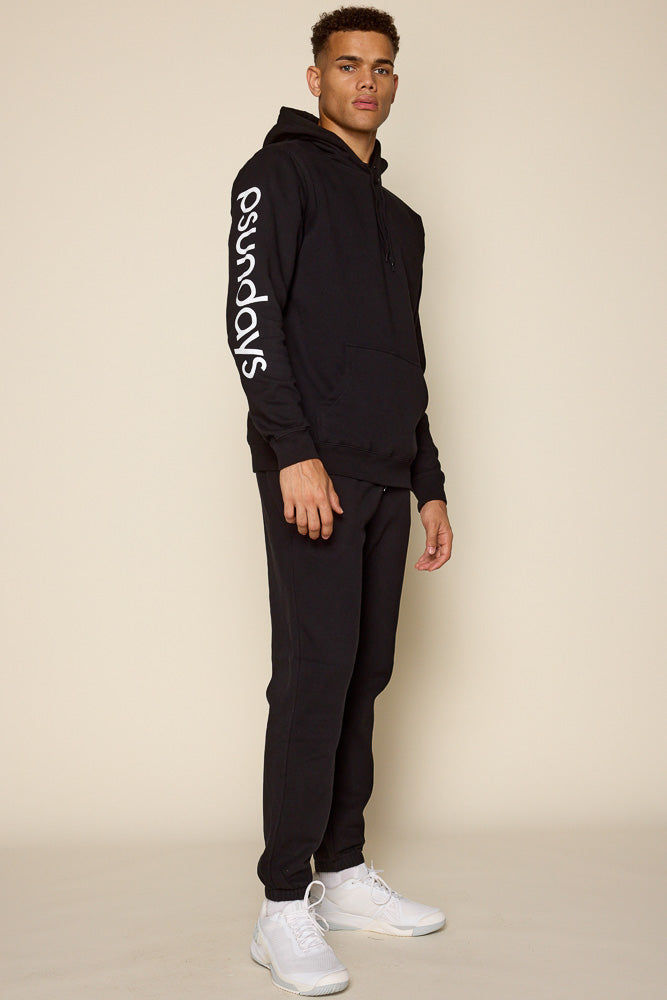 Full view of male model wearing a black hoodie with Psundays logo in white print along right sleeve. Model is also wearing black joggers and white sneakers to complete the look